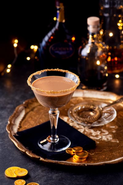 Hanukkah inspired chocolate martini on a gold tray with a bronze cocktail strainer and gelt. The glass in on a fringed black napkin.